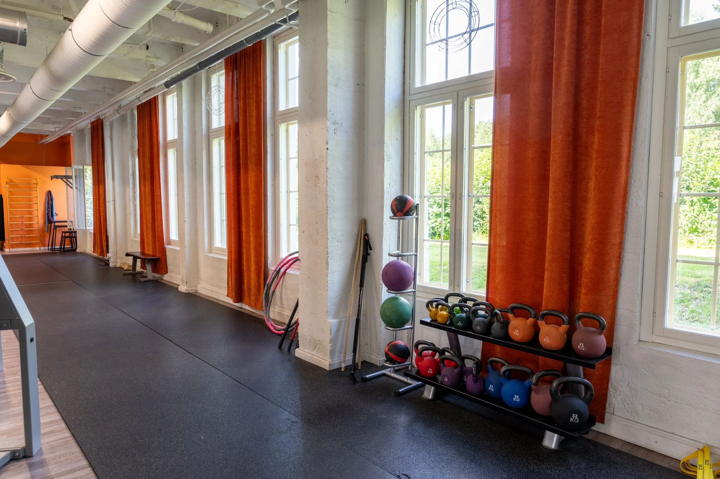 Kuntosalitila, jossa on käsipainoja, voimapalloja, hyppynauhoja ja punaisia kettlebell-levyjä, sekä suuri ikkuna ja oransseja verhoja.