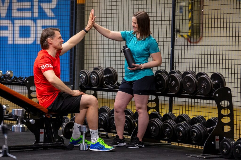 Personal trainer ja asiakas antavat high five kuntosalilla, painonnostovälineet taustalla.