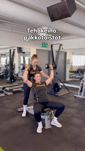 Kaksi naista treenaa painonnostossa salilla, toinen auttaa toista.