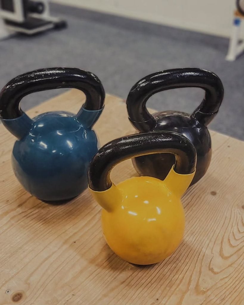Kolme eriväristä kettlebellia kuntosalilla, sininen, musta ja keltainen.